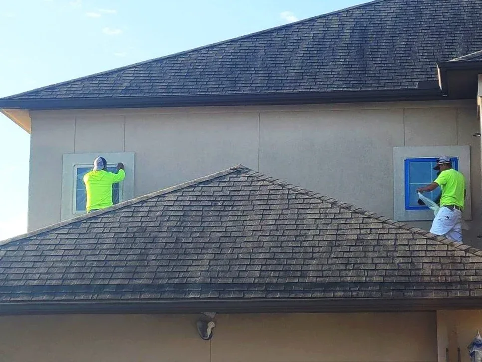 Two workers in neon shirts are installing or repairing corner window trim on the exterior of a beige house with a sloped shingled roof during daytime.
