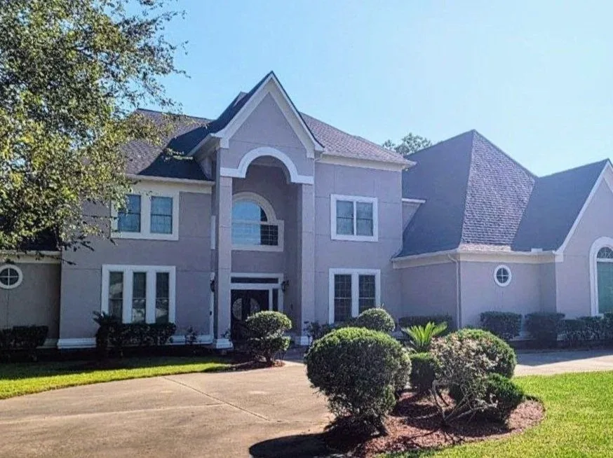 A large, gray two-story house with a decorative front entrance, multiple windows, and a well-maintained lawn with bushes and a tree.
