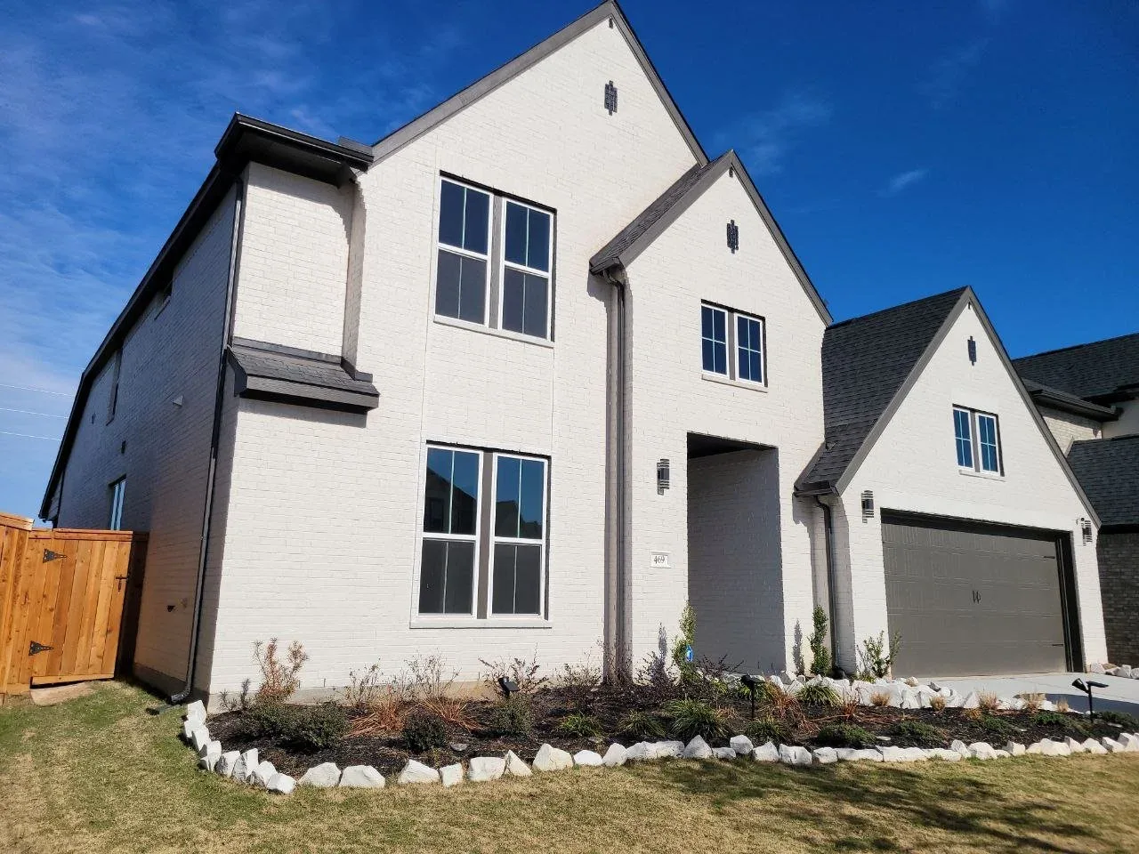 Modern white two-story house with black shingle roof and attached garage, front lawn with plants, under a blue sky.