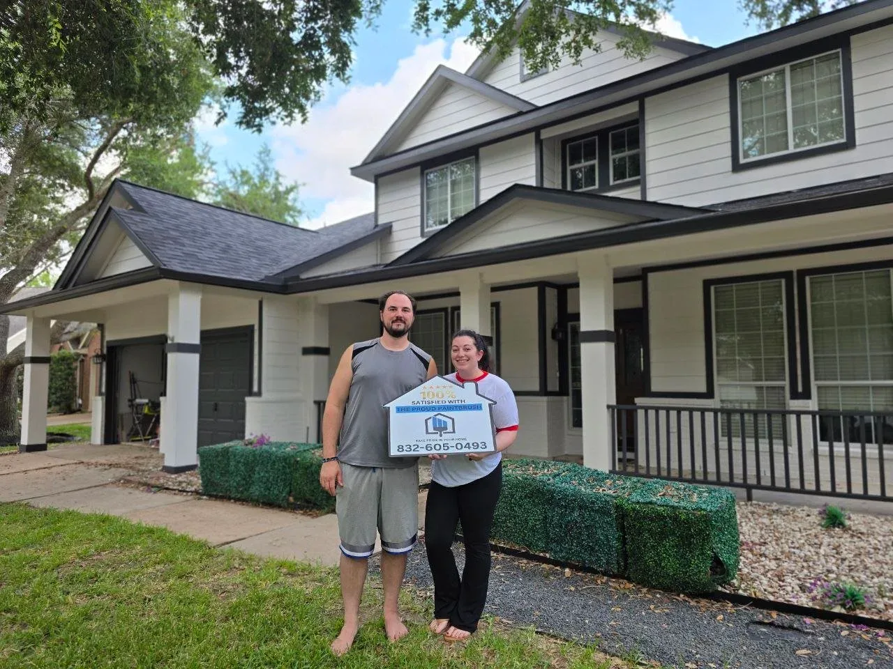Two people standing in front of a house, holding a sign that reads, '100% satisfied with the proud painter, 832-605-0493.' The house is large, white with black trim, and has a manicured lawn and a hedge in the front yard in richmond texas