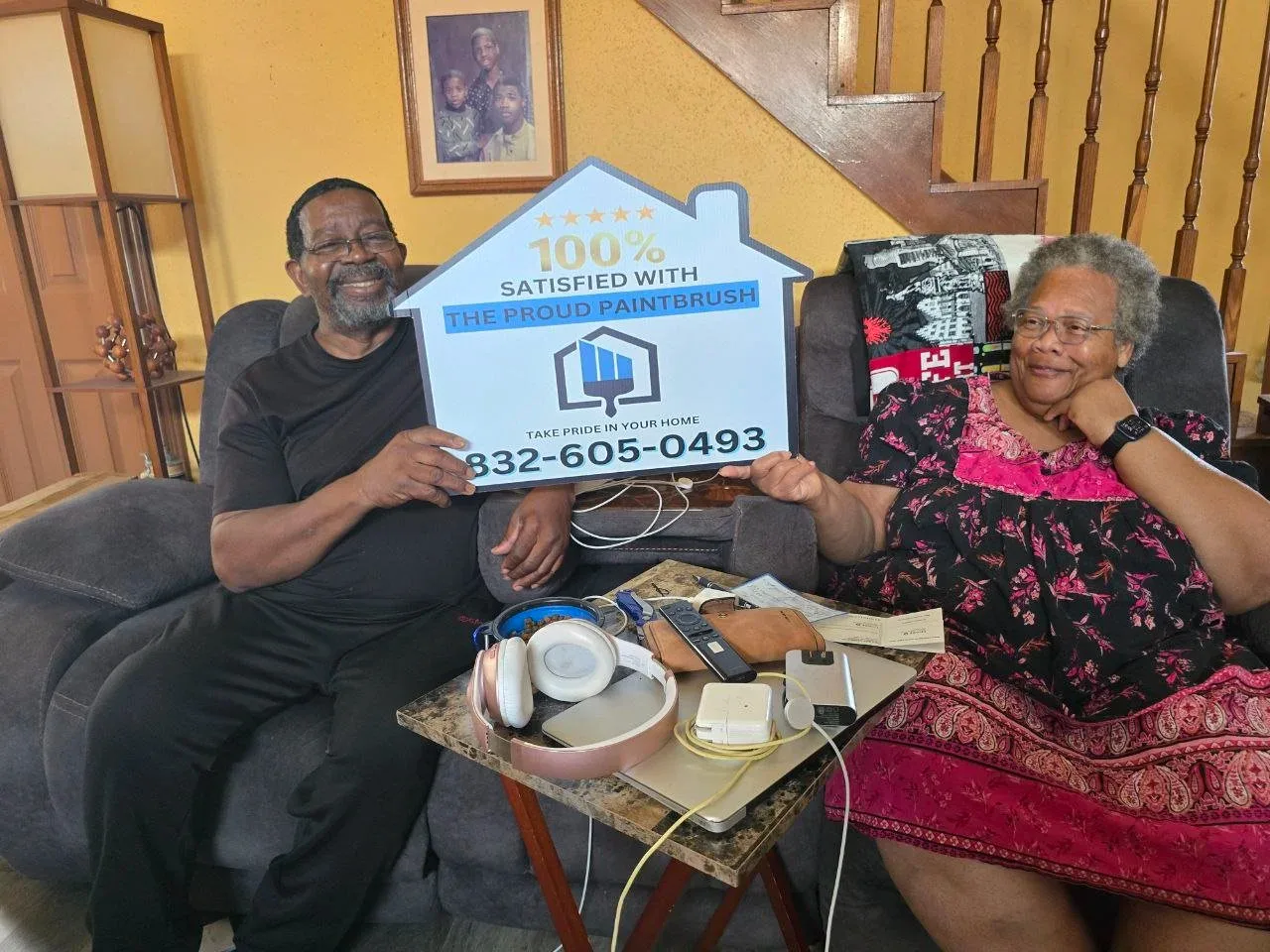 Two elderly people sitting on a couch, holding a large sign that reads, "100% satisfied with the proud paintbrush," with a phone number. They appear happy, with a man on the left smiling and a woman on the right resting her chin on her hand, smiling. A small table in front of them holds various electronic devices and accessories, and there is a photograph of children on the wall behind them.