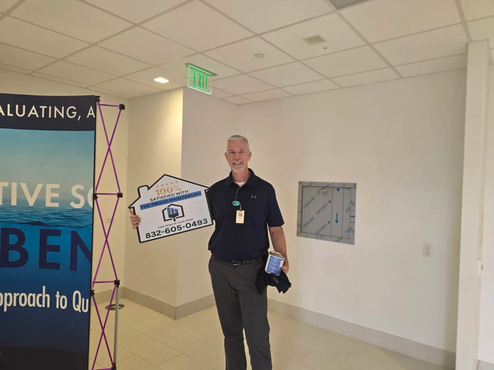 A man standing in a hallway holding a sign that says '100% satisfied with the proud paintbrush,' with a phone number. He is wearing a black shirt with a company logo and a name badge, and is smiling at the camera.